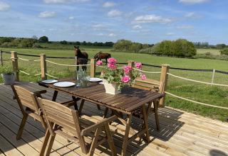 Wooden deck with a table and chairs, fresh flowers, and a scenic view of horses in a green UK field.