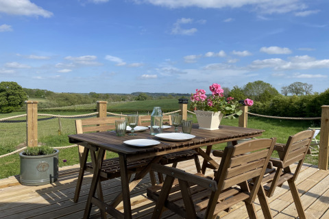 Wooden terrace with set dining table, flowers, and countryside view at The Campion pod in the United Kingdom.