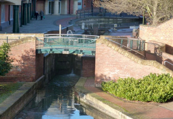 Photo d'une écluse de canal tranquille avec un pont en zone urbaine, entourée de murs en brique et de buissons verts.