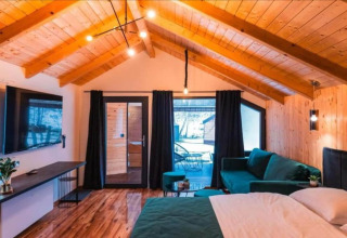 Chambre de chalet moderne avec plafond en bois, lit double, canapé vert, TV et accès à la terrasse.