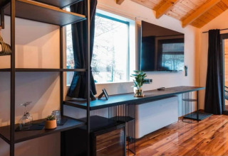 Intérieur de chalet moderne avec bureau noir, téléviseur mural, grande fenêtre, rideaux noirs et parquet.
