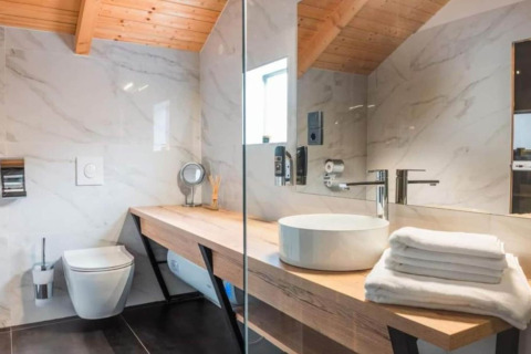 Salle de bain moderne avec plafond en bois, vasque, miroir, serviettes blanches et WC mural dans un chalet.