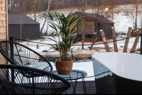 Terrasse extérieure de chalet avec chaise noire, plante, petite table et baignoire, vue sur paysage enneigé.