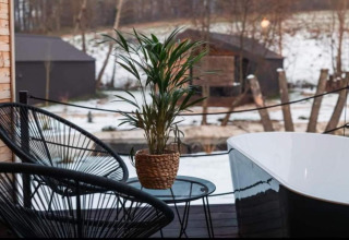 Terrasse extérieure de chalet avec chaise noire, plante, petite table et baignoire, vue sur paysage enneigé.