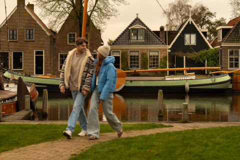 Stel wandelt hand in hand bij MarinaPark Beach Resort Soal in Friesland, Nederland, met boten en huizen op de achtergrond.