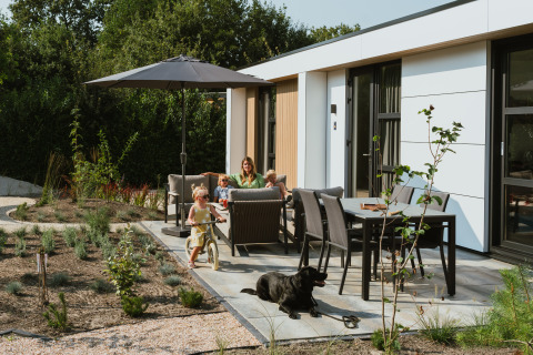 Familie nyder solrig terrasse med parasol, børn leger og sort hund slapper af, MarinaPark Residentie, Holland.