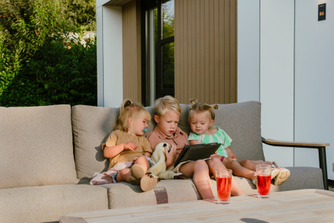 Three young children sit together on an outdoor sofa at MarinaPark Residentie Bloemendaal, looking at a tablet.
