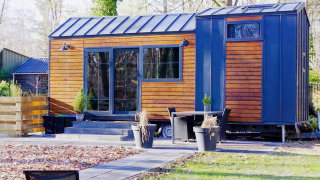 Charming Tiny House med træfacade og blå detaljer, omgivet af natur og moderne udendørsmøbler.
