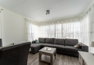Woonkamer met ruime grijze hoekzetel, salontafel en lichte gordijnen in luxe chalet ‘54 Bantega’ in Nederland.