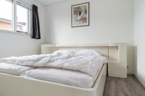 Heldere slaapkamer met tweepersoonsbed, witte muren, raam, donker gordijn en schilderij aan de muur.