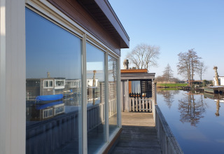 Vue du houseboat Luxury chalet '54 Bantega' aux Pays-Bas, eau calme, arbres et ciel bleu en arrière-plan.