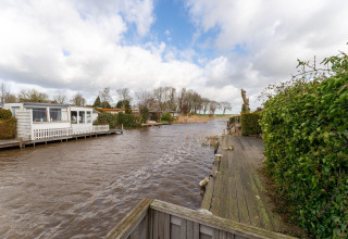 Husbaad ved vandkant med træterrasse og grøn hæk i Bantega, Holland, under en overskyet himmel.