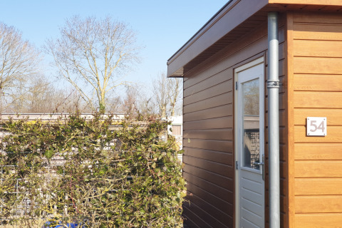 Buitenzicht op een modern houten huis met nummer 54 in het zonlicht en omringd door struiken en bomen.