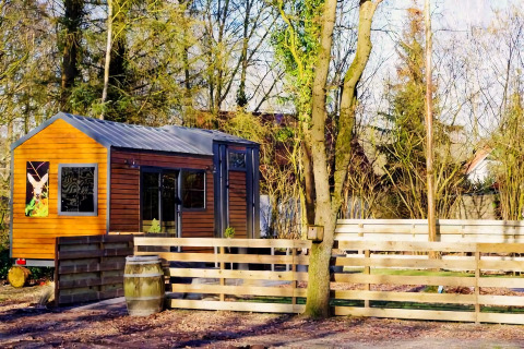 Charming Tiny House met houten gevel en hek, omringd door bomen in een rustige omgeving in Nederland.