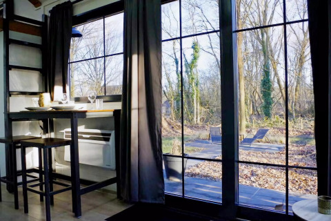 Vista desde el interior del Charming Tiny House en Países Bajos, con mesa, sillas y ventanales al bosque.