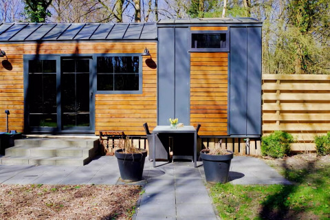 Charming Tiny House en Países Bajos con fachada de madera, terraza, mesa al aire libre y plantas verdes.