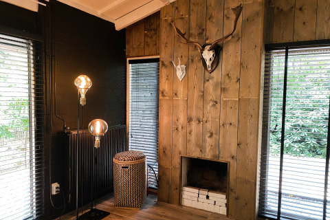 Woonkamer in Dutch Cabin Houses C66, Nederland, met houten wand, open haard, gewei en grote ramen.