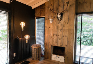 Woonkamer in Dutch Cabin Houses C66, Nederland, met houten wand, open haard, gewei en grote ramen.