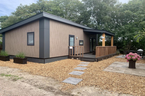 Modern chalet met terras en veranda, omgeven door bomen en planten, met een stenen looppad ervoor.