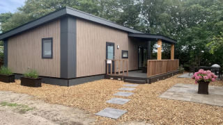 Moderne chalet med terrasse og veranda, omgivet af træer og blomsterkrukker, udendørs sti.