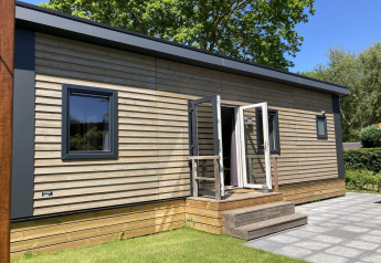 Chalet moderno con rivestimento in legno, giardino e terrazza, porte aperte e alberi sullo sfondo.