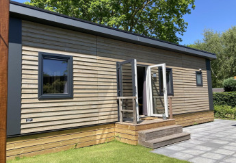 Moderne chalet med træbeklædning, have og terrasse, solrigt og omgivet af grønne træer og græsplæne.