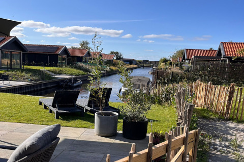 Udsigt fra terrasse med liggestole og planter ved kanal og hytter på en solrig dag i Holland.