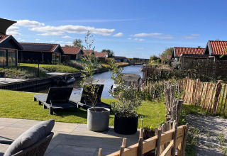 Udsigt fra terrasse med liggestole og planter ved kanal og hytter på en solrig dag i Holland.