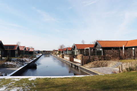 Scenic Stay with hytter ved en kanalside i Holland, omgivet af sne og grønne områder under blå himmel.