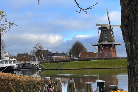 Molino de viento junto al canal con botes, árboles y casas al fondo, en Scenic Stay with en los Países Bajos.