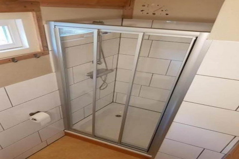Ducha con puertas de cristal y paredes de baldosas blancas en baño, rollo de papel higiénico en la pared