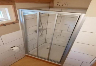 Brusekabine med glasdøre og hvid flisevæg i badeværelset, rulle toiletpapir på væggen, naturligt lys