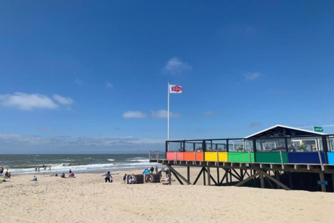 Casa de playa con barandilla de colores arcoíris y gente en la arena bajo un cielo azul claro.