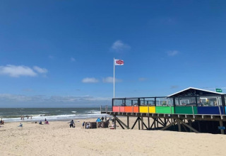 Casa de playa con barandilla de colores arcoíris y gente en la arena bajo un cielo azul claro.