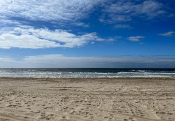 Strandudsigt fra et strandhus med sand, havbølger og en blå himmel med lette skyer en rolig dag.