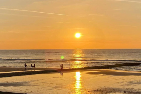 Zonsondergang aan zee gezien vanuit een strandhuis, met mensen die wandelen en kinderen spelend op het strand.