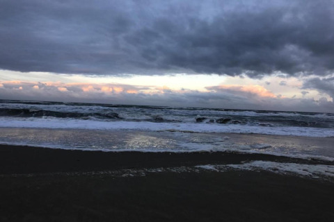 Vista de una playa tormentosa con nubes oscuras, olas espumosas y débil luz solar en el horizonte.