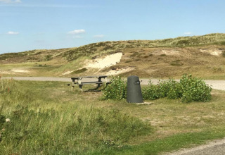 Et strandlandskab med bord-bænkesæt, skraldespand og klitter i baggrunden under en klar himmel.