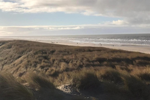 Uitzicht vanuit een strandhuis met grasduinen, zandstrand en enkele mensen langs de zee onder bewolkte lucht.