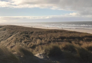 Vista desde una casa de playa con dunas cubiertas de césped, playa arenosa y algunas personas junto al mar.