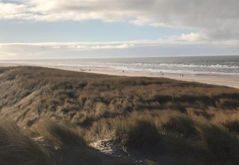 Blick von einem Strandhaus auf grasbewachsene Dünen, weitläufigen Strand und Menschen am Meer.