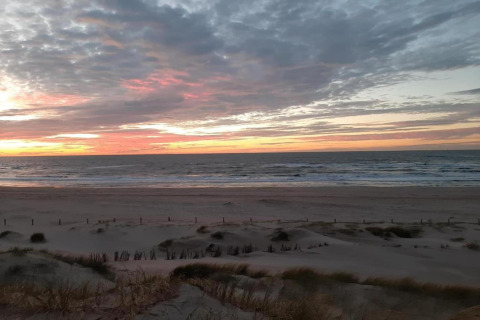 Coucher de soleil sur la plage vu depuis une maison au bord de mer, avec dunes, vagues et ciel nuageux.