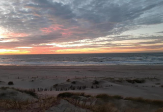 Solnedgang ved stranden set fra et strandhus, med skyer og bølger, omgivet af klitter og marehalm.