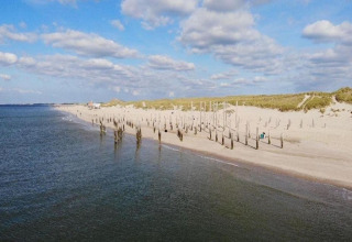 Strandzicht met houten palen in het zand, nabij de zee, onder een deels bewolkte blauwe hemel.