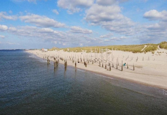 Strandansicht mit Holzpfählen im Sand und ruhigem Meer daneben unter einem bewölkten Himmel.