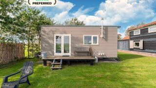 Tiny House moderne avec terrasse en bois et chaise sur pelouse verte sous un ciel bleu et nuageux.