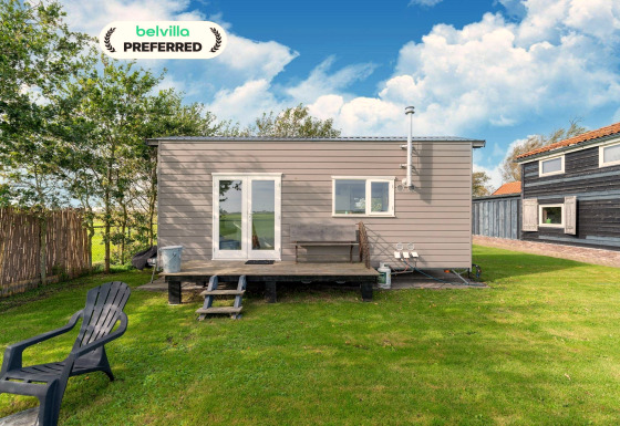 Kleine moderne Tiny House met terras, kunststof stoel en grasveld onder een blauwe lucht met wolken.