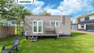 Lille moderne Tiny House med trappe, grøn græsplæne og plastikstol under blå himmel og skyer.