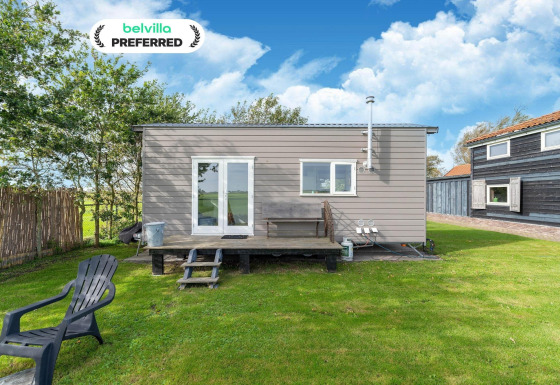 Pequeña casa Tiny House con porche de madera, césped verde y silla bajo un cielo azul y nubes.