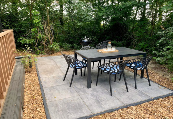 Buiten eethoek bij een lodge, zwart tuinmeubilair op betegeld terras, omringd door groene bomen.